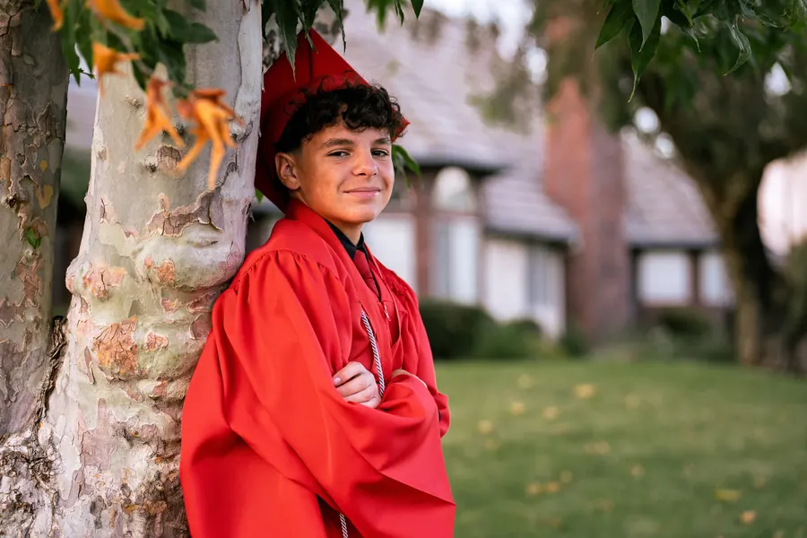Young graduate in a red gown and cap with arms crossed by a tree
