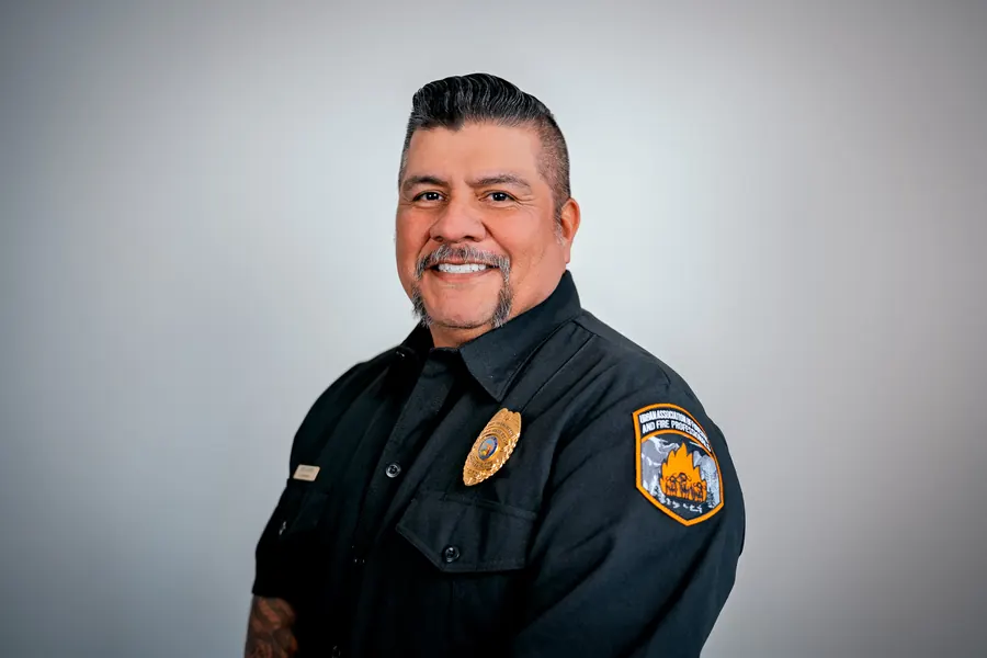 Smiling fire service professional in a dark uniform with a badge, studio headshot on a gray backdrop