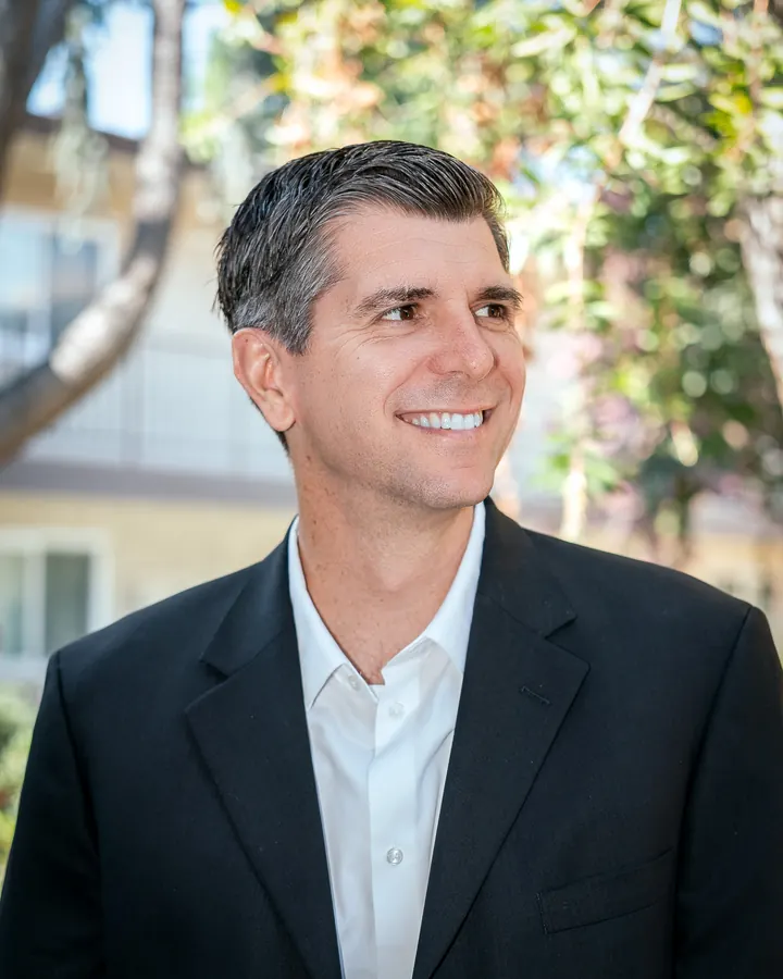 Man in a dark blazer smiling outdoors under trees