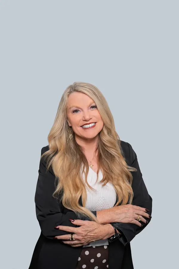 Smiling woman with long blonde hair and crossed arms, studio headshot on a light backdrop