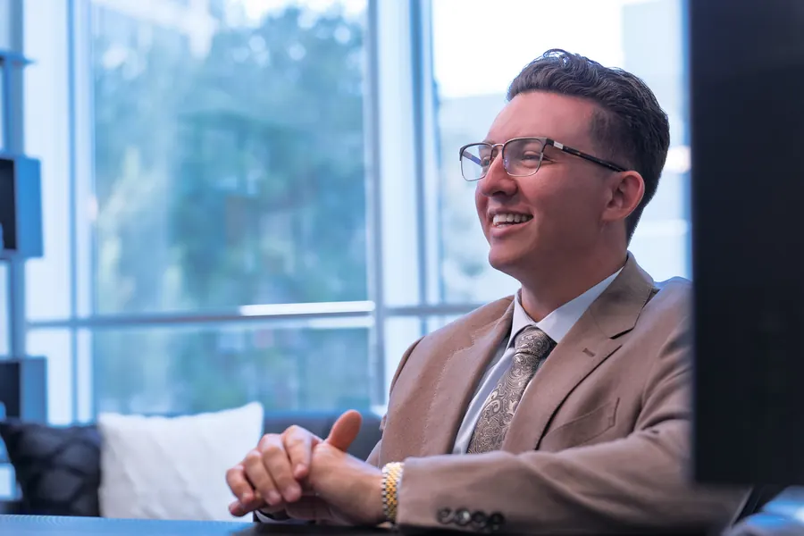 Smiling man in a tan suit seated at a desk by bright windows